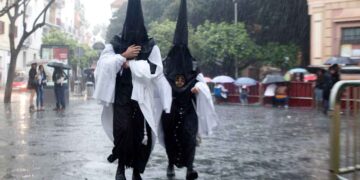Dos nazarenos con capirotes negros caminan bajo la lluvia en la Semana Santa de Andalucía